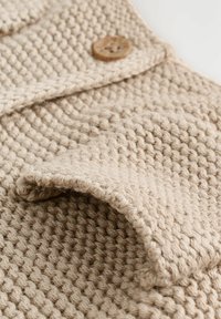 Tissu tissé beige avec un motif texturé, mettant en avant un détail de bouton. Texture douce et chunky avec un col replié et des coutures lisses.