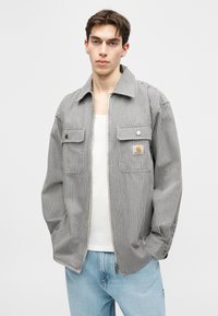 Noor mees, kes kannab halli ja valget vertikaaltriibulist lukuga jopet valge tanktopi ja helesiniste teksade kohal, seisab käed taskutes.