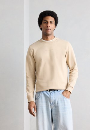 Young man with curly hair wearing a beige sweatshirt and light blue jeans, standing with one hand in pocket against plain wall.