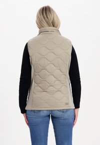 Beige gesteppte Weste mit hohem Kragen, kombiniert mit einem schwarzen Langarmtop und blauen Jeans. Die Weste zeichnet sich durch eine glatte Textur und keine sichtbaren Beschläge aus.