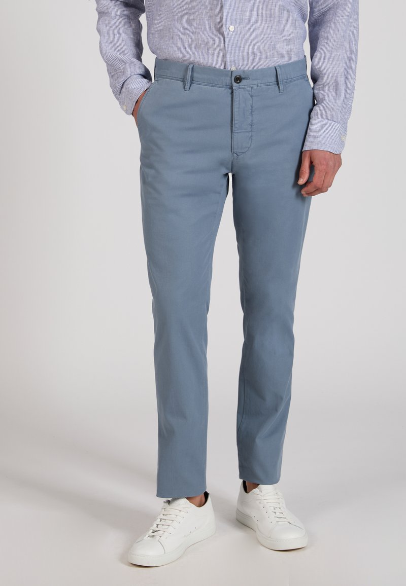 Man wearing light blue trousers, a blue striped button-up shirt, and white sneakers, standing with one hand in pocket.