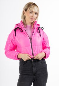 Ružová softshellová bunda s kapucňou, trblietavý povrch, čierne šnúrky a predný zips. Skombinovaná s svetloružovým topom a čiernymi džínsy.