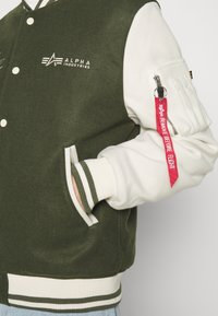 Giacca varsity verde e bianca con polsini a coste. Presenta un logo, tasche laterali e un'etichetta rossa "RIMUOVERE PRIMA DEL VOLO" sulla manica.