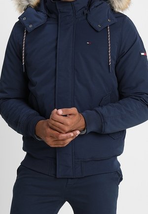 Homme portant une veste d'hiver bleu marine avec capuche doublée de fourrure, poches avant et petits écussons logo sur la poitrine et le bras, mains jointes devant.