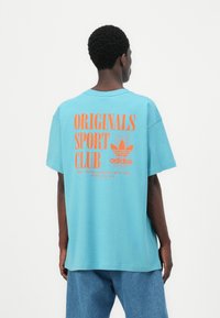 Modra bombažna majica z izrazitim oranžnim napisom "ORIGINALS SPORT CLUB" in logotipom Adidas na hrbtni strani. Ohlapen kroj, kratke rokave.