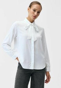 Biała bluzka z kołnierzykiem i detalem w postaci kokardy, długimi rękawami oraz gładkim, lekkim materiałem. Noszona do ciemnoszarych dżinsów.