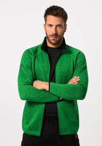 Chaqueta verde con cremallera y tejido texturizado, con detalles en negro, cuello alto y mangas ajustadas, llevada sobre una camisa negra y combinada con pantalones oscuros.