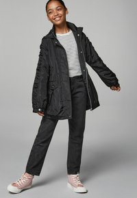 Giacca nera in nylon con cappuccio e maniche arricciate, indossata sopra una maglietta a righe. Abbinata a jeans neri a vita alta e sneakers rosa.
