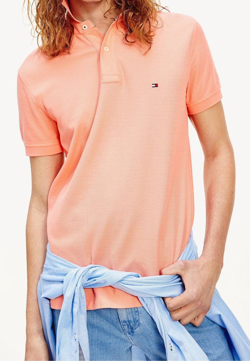 Kortärmad polotröja i korall med klassisk krage, med en liten logotyp på bröstet och en ljusblå skjorta knuten runt midjan.
