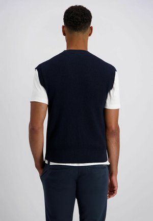 Homme portant un gilet sans manches en tricot bleu marine sur une chemise blanche à manches courtes et un pantalon bleu marine, vu de dos avec les mains dans les poches.