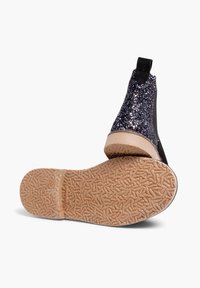 Botín negro hasta el tobillo con parte superior de purpurina brillante, paneles laterales elásticos y suela de goma texturizada. La suela presenta un patrón ondulado para mayor agarre.