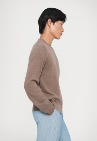 Brun strikket sweater med rund halsudskæring, lange ærmer og sideventiler, parret med lyseblå stribede bukser. Glat tekstur, tætsiddende design.