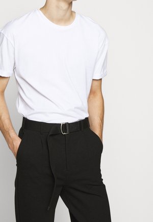 Personne portant un t-shirt blanc uni à manches courtes rentré dans un pantalon noir taille haute avec une ceinture noire, les mains dans les poches.