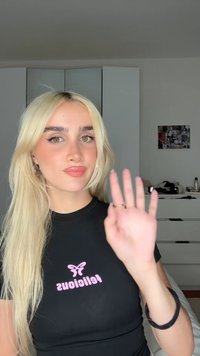 Svart t-shirt med rosa texten "felicious", långt blont hår och en hand höjd i en vinkning. Bakgrunden har enkel vit möblering.