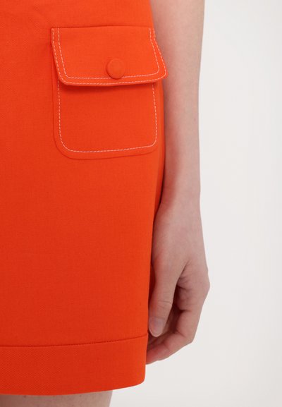 Oranje rok met een heupzak voorzien van een afgeronde klep en knop. De stof heeft een gestructureerde afwerking en een gegolfde rand.