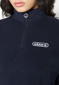 Närbild av person som bär en marinblå fleece-tröja med zip i kvartslång modell, med vit Adidas-logotyp och metalldragkedja med Adidas-emblem.