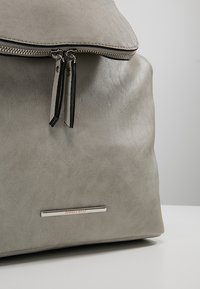 Sac à dos en cuir gris clair avec double curseur de fermeture éclair et plaque de marque en métal portant l’inscription ANNA FIELD sur la partie inférieure avant.