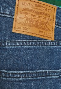 Papel em couro castanho da Levi's em jeans de denim azul, mostrando o modelo 502, cintura 31, comprimento 32, e o logótipo da marca com dois cavalos.