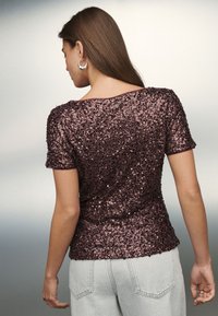 Bluza cu mânecă scurtă, de culoare mov închis, cu aplicații din paiete. Prezintă un decolteu rotund și un croi mulat, asortată cu pantaloni din denim gri deschis.