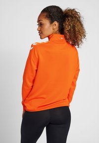 Giacca sportiva arancione con colletto alto, dettagli bianchi a motivo sulle spalle e un tessuto morbido e leggero. Leggings neri visibili.