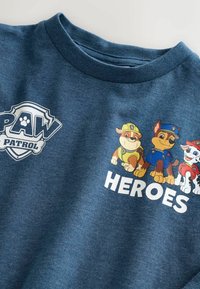 Blå skjorta med Paw Patrol-logotyp, tre tecknade hundar i räddningsuniformer och ordet "HEROES" i vit text.