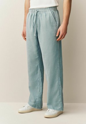 Muž oblečený v svetlomodrých ľanových nohaviciach s elastickým pásom a bočnými vreckami, doplnených bielymi teniskami a bielym topom.