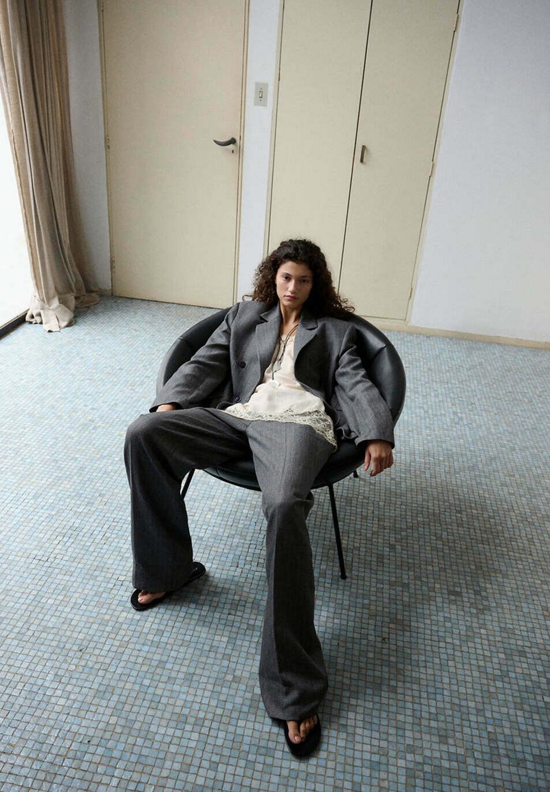 Femme aux cheveux bouclés portant un costume gris oversize et un chemisier en dentelle blanche, allongée dans une chaise noire sur un sol carrelé dans une pièce simple.