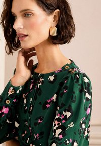 Groene blouse met een gedurfd patroon van zwarte, witte en roze vlekken. Heeft knoopdetails op de schouder en de manchet, gemaakt van een zachte stof.