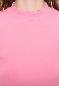 Κοντινό πλάνο ενός ατόμου που φοράει ροζ ριπ τοπ με κυματιστό ψηλό ντεκολτέ και μακριά καστανά μαλλιά ορατά στις πλευρές.