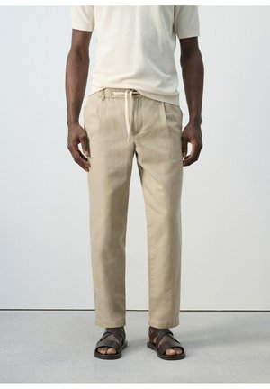 Mężczyzna w beżowych spodniach ze sznurkiem, białej koszuli z krótkim rękawem i ciemnobrązowych skórzanych sandałach stojący na jednolitym tle.