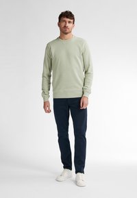Sudadera verde claro con puños y dobladillo acanalados, combinada con pantalones azul marino oscuros y zapatillas blancas, sobre una figura en un fondo liso.