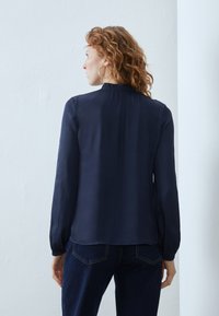 Blusa azul marino con cuello alto, mangas largas y detalles fruncidos en el cuello; textura suave y tejido ligero.