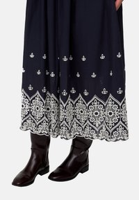 Dunkelblauer Rock mit aufwändiger weißer Blumenstickerei entlang des Saums, kombiniert mit braunen Lederstiefeln. Der Rock fällt locker von der Taille.