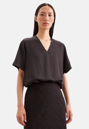 Vrouw met lang donker haar draagt een donkere blouse met V-hals en een hoog getailleerde rok met bloemenkant patroon op een lichte achtergrond.