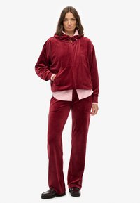 Ensemble en velours rouge composé d'une veste à capuche zippée et d'un pantalon. La veste présente un petit logo et une poche kangourou ; le pantalon est évasé.
