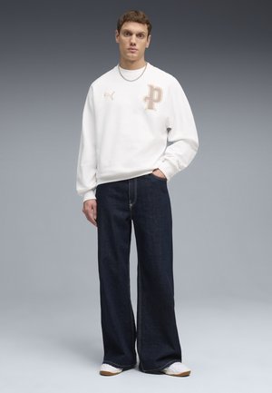 Junger Mann steht mit der Hand in der Tasche, trägt ein weißes Sweatshirt mit Puma-Logo, dunkle weite Jeans, weiße Sneakers und eine silberne Halskette.