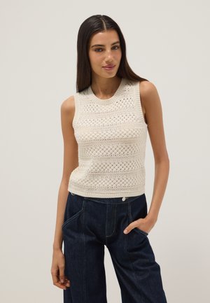 Donna con lunghi capelli scuri che indossa un top a maglia bianco senza maniche con motivi e jeans scuri a vita alta, in piedi con una mano in tasca.