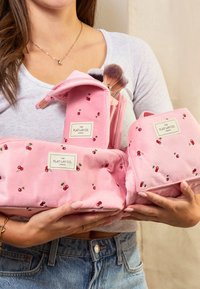 Roze stoffen make-up tassen met kersenborduring en merklabels, met een ritssluiting en een borstelhouder, vastgehouden in handen.