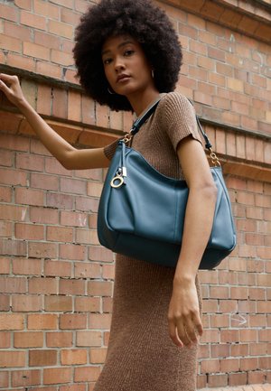 Kobieta o kręconych włosach w brązowej sukience, niosąca dużą niebieską skórzaną torebkę na tle ceglastej ściany