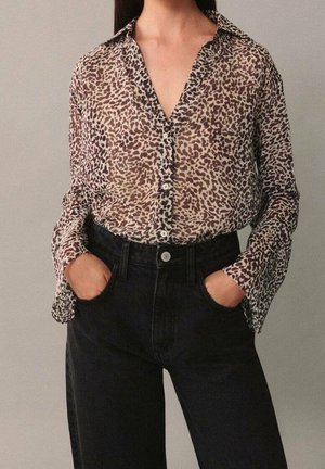Femme portant un chemisier transparent à manches longues imprimé animalier, boutonné, rentré dans un jean noir taille haute, mains dans les poches.