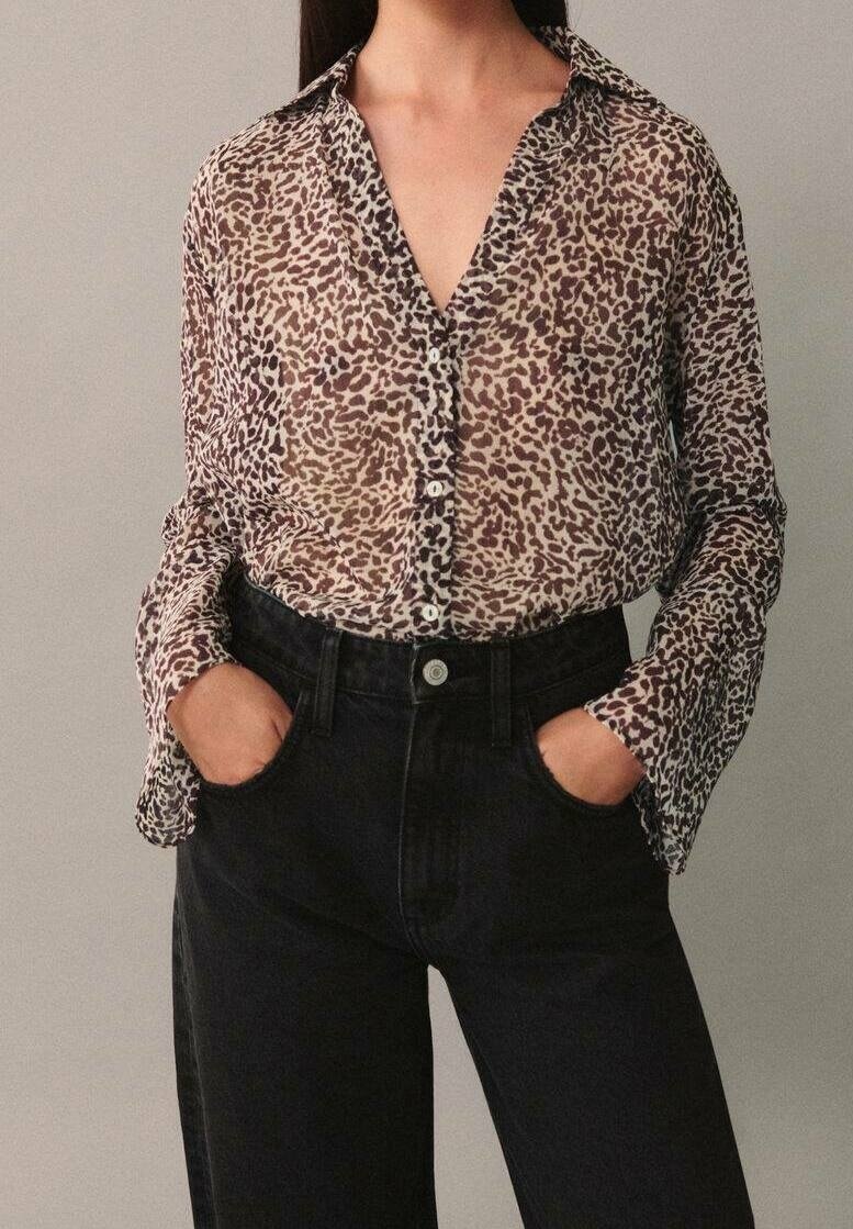 Femme portant un chemisier transparent à manches longues imprimé animalier, boutonné, rentré dans un jean noir taille haute, mains dans les poches.