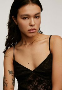 Camiseta de encaje negro con tirantes finos, con estampados florales. Se lleva un collar delicado con un pequeño colgante en forma de "B" alrededor del cuello.
