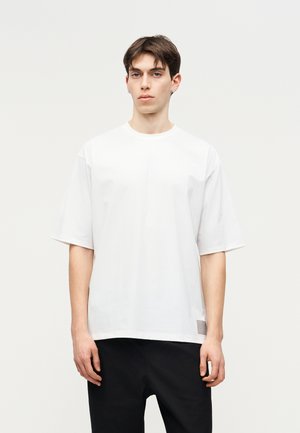 Fiatal férfi rövid sötét hajjal, sima fehér, túlméretezett pólót és fekete nadrágot visel, világos, semleges háttér előtt áll.