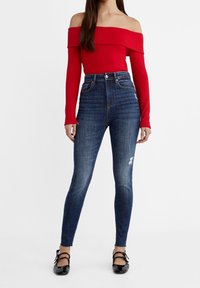 Top rojo acanalado con escote off-the-shoulder, combinado con jeans de tiro alto de color azul oscuro que presentan un detalle desgastado en el muslo. Zapatos negros completan el conjunto.