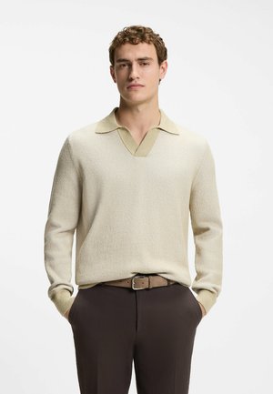 Jeune homme portant un pull en maille crème avec un col et un pantalon marron foncé, debout les mains dans les poches devant un fond blanc.