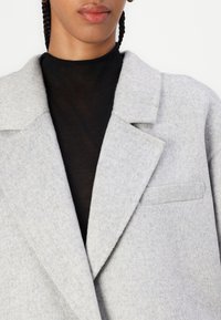 Blazer de mezcla de lana gris con solapas de muesca, líneas limpias y un único bolsillo en el pecho. Llevado sobre un suéter de cuello alto negro.