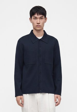 Jeune homme aux cheveux noirs courts portant une chemise bleu marine foncé boutonnée avec deux poches poitrine et un pantalon blanc sur un fond clair uni.
