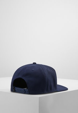 Casquette snapback bleu marine avec sangle réglable, six œillets de ventilation et visière incurvée, posée sur une surface blanche.