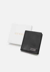 Portafoglio in pelle nera con logo argento, esposto aperto accanto a una scatola bianca con la scritta "GUESS" in lettere dorate. Texture liscia, design compatto.