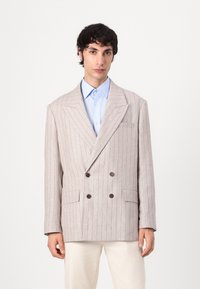 Double-breasted blazer in light beige fabric with subtle blue stripes, featuring large brown buttons and two front pockets, notched lapels.